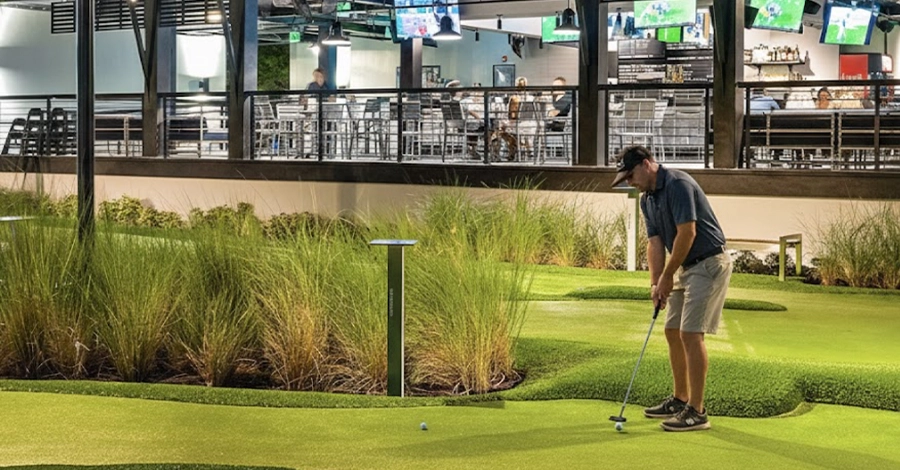 Man putting golf ball on mini golf green outside restaurant patio.