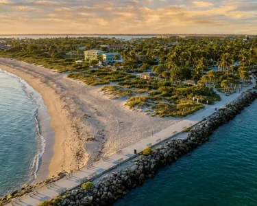 Aerial sunset view of a Florida coastal peninsula with white sand beach, rocky jetty walkway, and lush tropical homes nestled between calm turquoise waters.